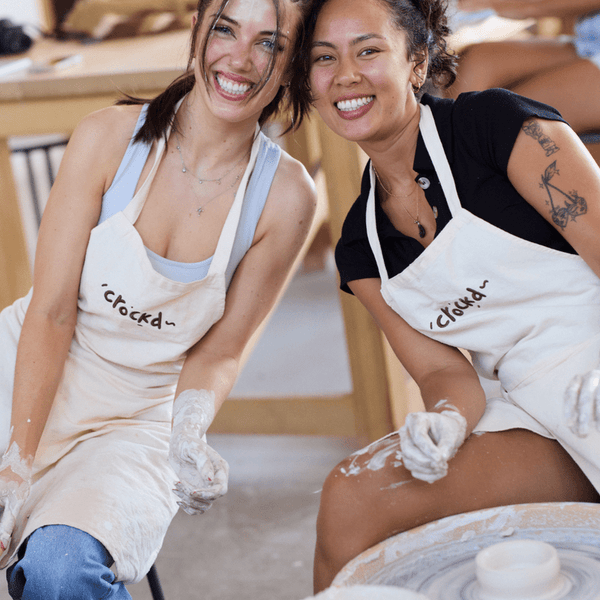 Smiling friends wearing aprons and working with clay at a beginner wheel throwing pottery class at Crockd Studios.
