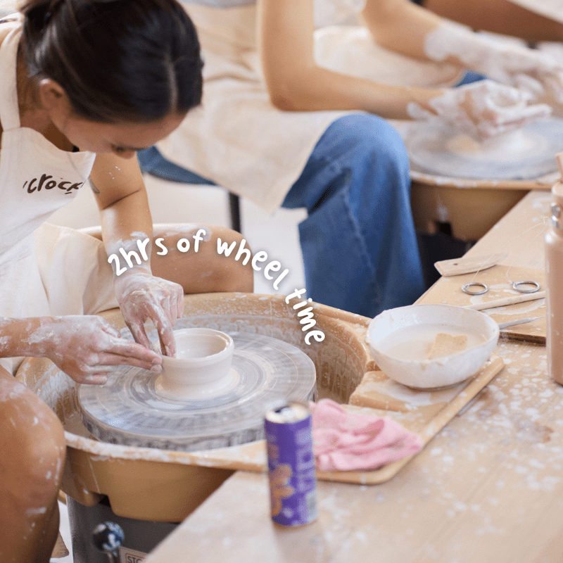 Smiling friends wearing aprons and working with clay at a beginner wheel throwing pottery class at Crockd Studios.